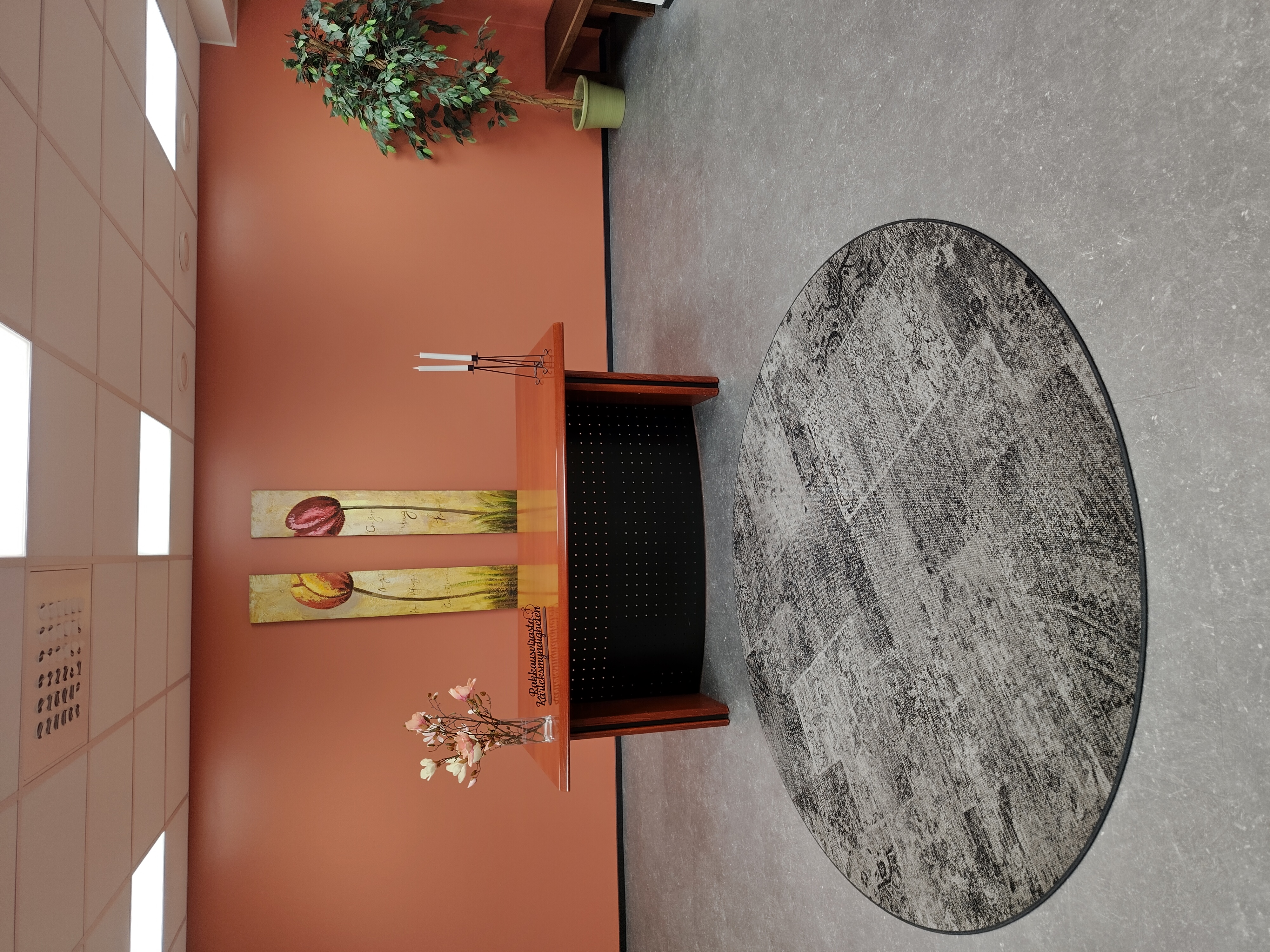 Digi- ja väestötietoviraston Oulun vihkitila. Vihkitilassa on pöytä, jonka edessä on suuri, pyöreä matto. Vihkitilan seinällä on kaksi taulua, jotka esittävät kukkia, jotka taipuvat toisiaan kohti.