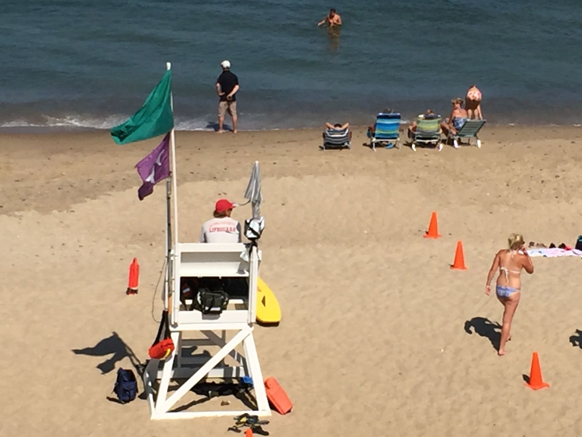 Rantavahti istuu tuolissa ja vartioi uimarantaa. Ihmiset ottavat aurinkoa rannalla tuoleissa. Yksi ihminen ui meressä.